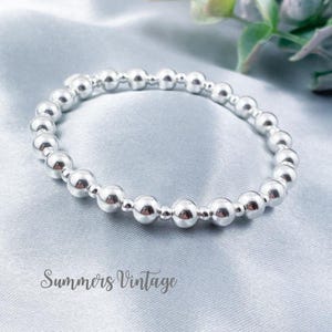 May include: A silver beaded bracelet with an elasticated design. The bracelet features multiple small, round, shiny beads. The background is a light blue, silky fabric. The text "Summers Vintage" is at the bottom.