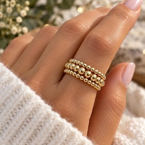 May include: A gold ring featuring three stacked bands of polished spherical beads. The design consists of two outer bands with smaller beads and a central band with larger beads, creating a uniform, rounded texture across the entire surface.