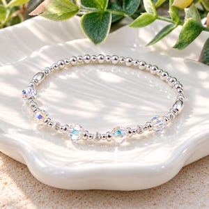 May include: A beaded bracelet featuring small, polished silver-toned spheres interspersed with larger, faceted clear crystal beads that display an iridescent, rainbow-like sheen. The design includes small, ribbed silver-toned accent beads.