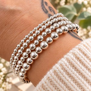 May include: A stack of four silver-toned beaded bracelets featuring polished, spherical beads of varying sizes. The bracelets are arranged in a row, showcasing a reflective metallic finish on each individual bead, creating a uniform and layered appearance.