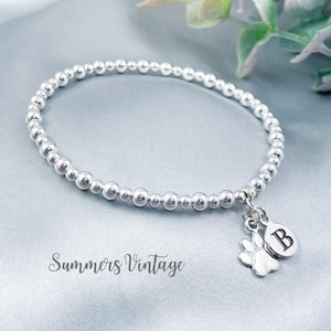 May include: Silver beaded bracelet featuring a paw print charm and a disc charm with the letter "B". The bracelet is displayed on a light blue surface. The words "Summers Vintage" are visible.