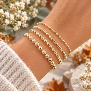 May include: A set of four gold-toned beaded bracelets worn together. Each bracelet features a different size of polished spherical beads, ranging from very small beads on the top strand to larger, uniform beads on the bottom strand.