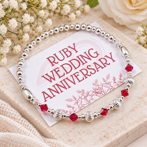May include: A circular beaded bracelet featuring polished silver-tone spheres, ribbed silver-tone accents, and faceted deep red crystal beads. The bracelet rests on a white card printed with the red text RUBY WEDDING ANNIVERSARY and floral line art.