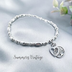 May include: A silver bracelet with a charm of a tree inside a heart. The bracelet is made of small silver beads.