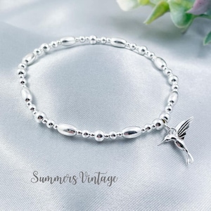 May include: Silver beaded bracelet with a hummingbird charm. The bracelet features small, round beads and larger, oval-shaped beads. A detailed hummingbird charm hangs from the bracelet. The text "Summers Vintage" is at the bottom.