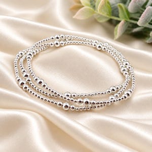 May include: A multi-strand bracelet composed of polished silver-toned beads of varying sizes. The design features a repeating pattern of smaller spherical beads interspersed with larger, prominent silver-toned spheres arranged in a circular loop.