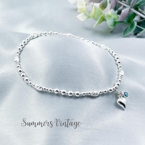 May include: A silver beaded anklet with a small heart charm and a light blue gemstone. The anklet is on a white satin background.