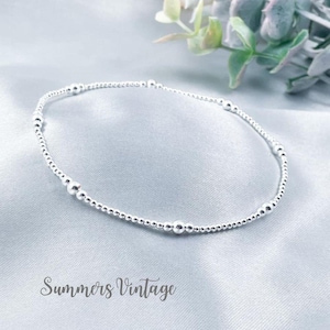 May include: A delicate silver bracelet with small beads and larger beads spaced out along the chain. The bracelet is on a light grey fabric background.