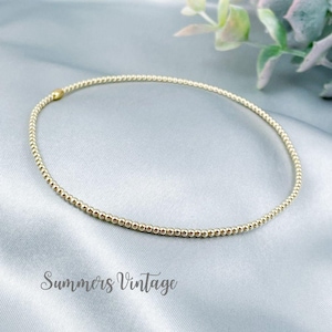May include: A gold beaded bracelet with a delicate design. The bracelet is made of small, round beads strung together to form a flexible band. The bracelet is displayed on a light blue surface. The text "Summers Vintage" is at the bottom.