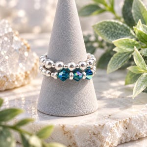 May include: A stackable ring featuring two bands of small, polished silver-toned beads. The lower band is accented with three faceted, iridescent teal-blue crystal beads that catch the light, creating a vibrant contrast against the metallic silver finish.