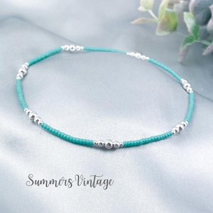 May include: A turquoise beaded anklet with silver accent beads. The anklet is displayed on a light blue satin surface. The text "Summers Vintage" is at the bottom of the image.
