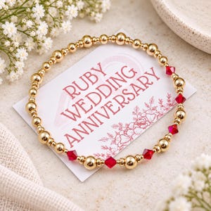May include: A circular stretch bracelet featuring polished gold-tone beads interspersed with faceted red crystal beads. The bracelet rests on a white rectangular card printed with the text RUBY WEDDING ANNIVERSARY in red, accompanied by a floral line art design.