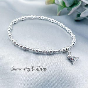 May include: A silver beaded bracelet with a small silver bird charm. The bracelet is on a grey background.