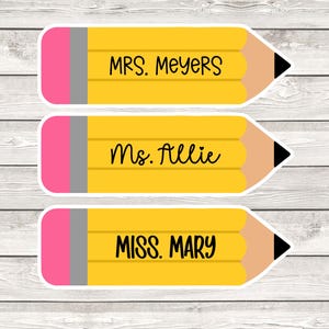 May include: Three pencil-shaped stickers with names: "MRS. MEYERS", "Ms. Allie", and "MISS. MARY". Each sticker has a yellow body, pink eraser, gray ferrule, and black tip. The background is a light wood grain.