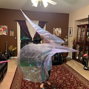 May include: A person is holding and twirling a large iridescent white fabric in a room with a brown wall and a patterned rug.