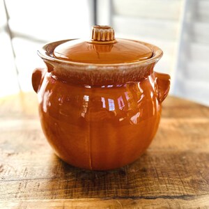 May include: A glazed ceramic casserole dish with a lid. The dish is a warm, earthy orange-brown color with a lighter rim and a small handle on the lid. It has two side handles and sits on a wooden surface.