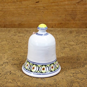 May include: A white ceramic bell with a blue and yellow geometric pattern. The bell has a rounded top and a wide base.