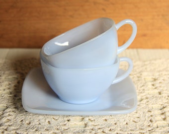 May include: Two light blue teacups stacked on a matching saucer. The cups have a rounded shape with a handle. The saucer is square with rounded corners. The set is on a cream-colored, crocheted surface.