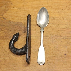 May include: A black metal hook with a pointed tip and a silver spoon on a wooden surface.