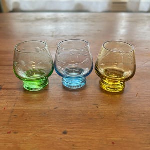 May include: Three small, stemless glasses in shades of green, blue, and yellow. Each glass has a clear bowl with etched leaf designs and a colored base. The glasses are arranged on a wooden surface.