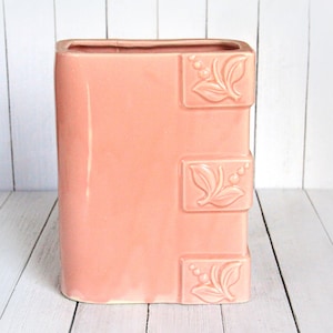 May include: A pink ceramic book-shaped planter with a floral design. The planter has a rectangular shape and is decorated with three raised floral motifs on the side.
