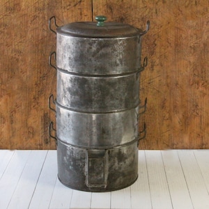 May include: Vintage metal steamer with four stacked compartments. The steamer has a dark, weathered appearance with handles on each section and a green knob on the lid. It sits on a white surface against a wooden background.