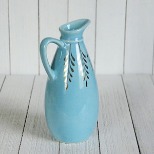 May include: A small, light blue ceramic pitcher with a gold leaf design. The pitcher has a rounded body and a handle.