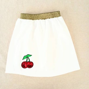 Peut inclure: Une jupe blanche avec une ceinture élastique dorée et une applique de cerise rouge.