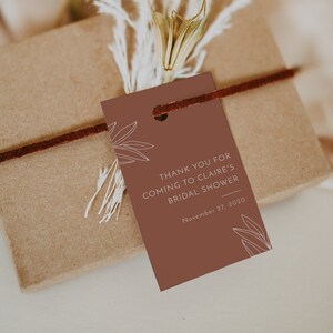May include: A gift box wrapped in brown paper, tied with a dark brown cord, and adorned with a terracotta-colored tag. The tag reads "THANK YOU FOR COMING TO CLAIRE'S BRIDAL SHOWER November 27, 2020". Dried flowers and foliage add a natural touch.