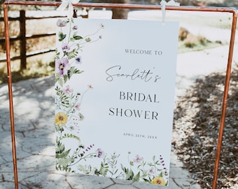 May include: A white rectangular sign suspended from a copper pipe frame. The sign features colorful watercolor floral illustrations along the left and bottom edges, with the text "WELCOME TO Scarlett's BRIDAL SHOWER APRIL 26TH, 20XX" printed in black.