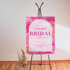 May include: A pink bridal shower sign on a black easel. The sign reads "Welcome to Taylor's Bridal Shower" with the date and countdown. A vase with dried flowers and a palm leaf is in the foreground.