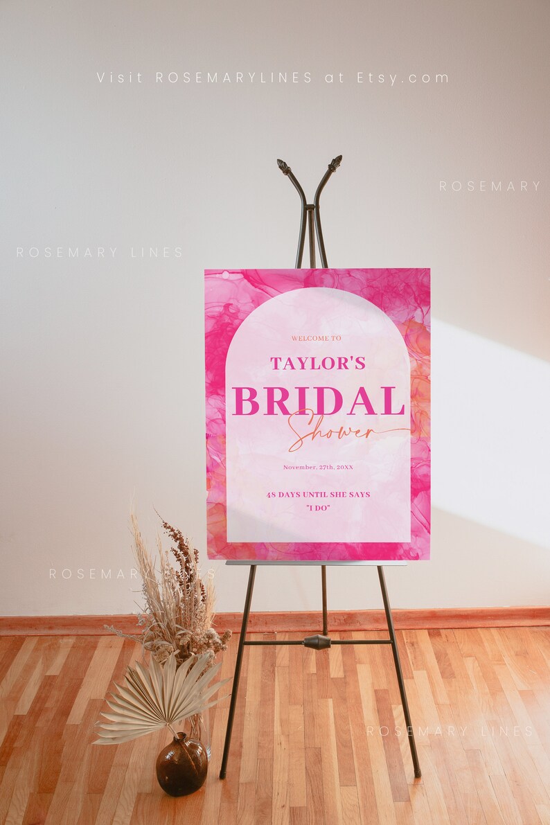 May include: A pink bridal shower sign on a black easel. The sign reads "Welcome to Taylor's Bridal Shower" with the date and countdown. A vase with dried flowers and a palm leaf is in the foreground.