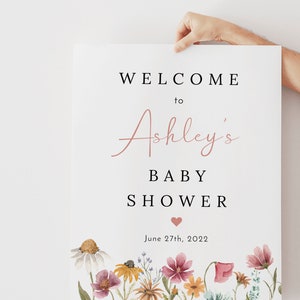 May include: A white sign with black text announcing a baby shower for Ashley. The text reads "Welcome to Ashley's Baby Shower" with a pink heart and the date "June 27th, 2022". The bottom of the sign features a watercolor illustration of a field of wildflowers in shades of pink, yellow, and blue.