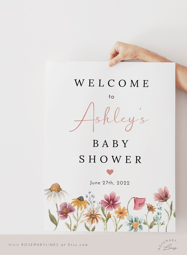 May include: A white sign with black text announcing a baby shower for Ashley. The text reads "Welcome to Ashley's Baby Shower" with a pink heart and the date "June 27th, 2022". The bottom of the sign features a watercolor illustration of a field of wildflowers in shades of pink, yellow, and blue.