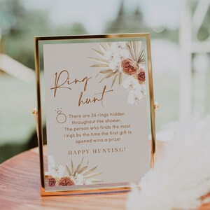 May include: A framed sign with the words "Ring Hunt" in elegant script, adorned with floral accents. The sign details a game with 24 hidden rings and the text "Happy Hunting!" The sign is in a gold frame.