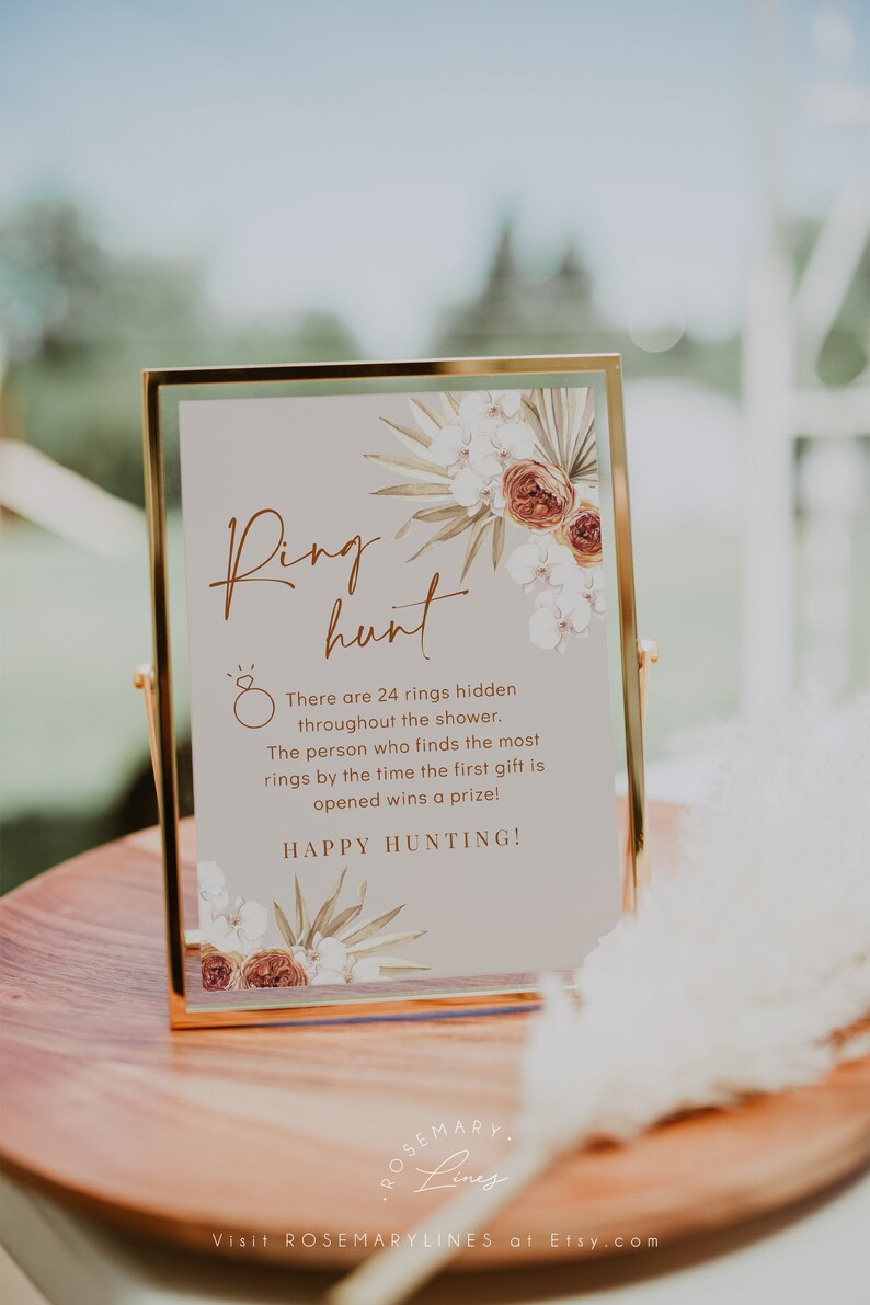 May include: A framed sign with the words "Ring Hunt" in elegant script, adorned with floral accents. The sign details a game with 24 hidden rings and the text "Happy Hunting!" The sign is in a gold frame.