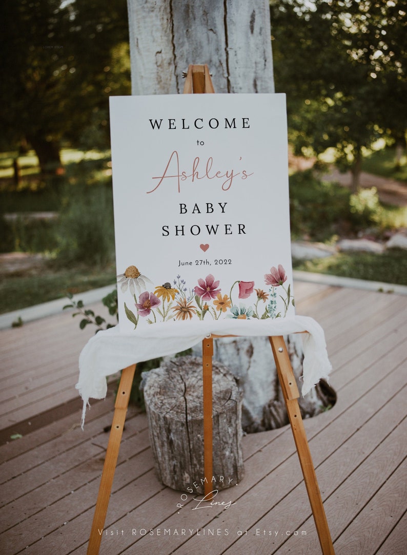 May include: A white sign with a floral border and the text "Welcome to Ashley's Baby Shower June 27th, 2022".