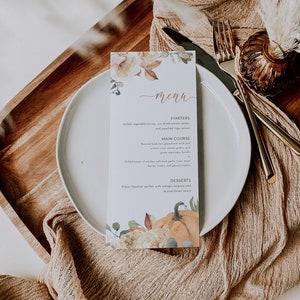 K&ouml;nnte beinhalten: Eine rechteckige Men&uuml;karte mit floralem und K&uuml;rbis-Design, mit dem Wort "Men&uuml;". Die Speisekarte listet Vorspeisen, Hauptgerichte und Desserts auf. Die Karte liegt auf einem wei&szlig;en Teller mit goldenem Besteck und einer beigen Leinen-Serviette.