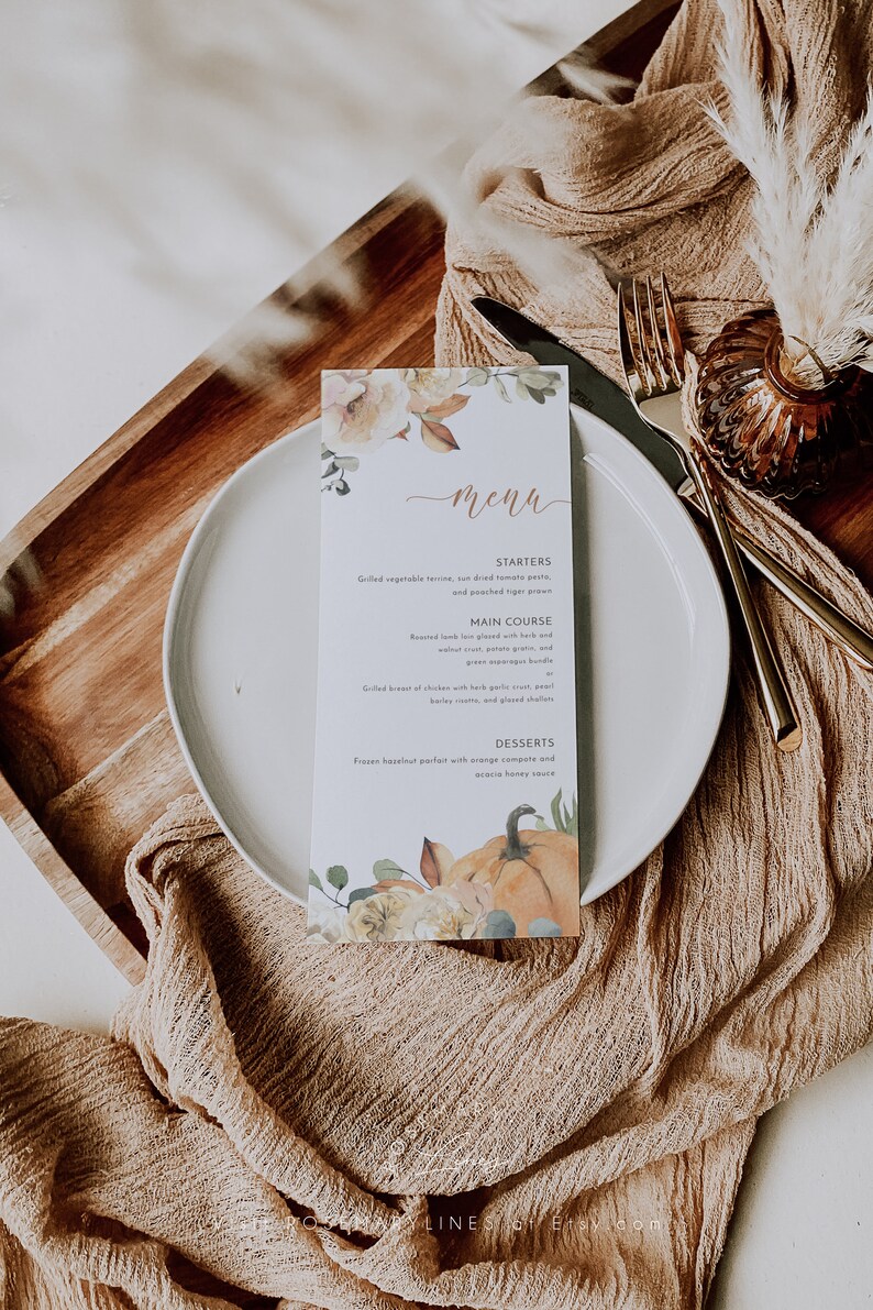 K&ouml;nnte beinhalten: Eine rechteckige Men&uuml;karte mit floralem und K&uuml;rbis-Design, mit dem Wort "Men&uuml;". Die Speisekarte listet Vorspeisen, Hauptgerichte und Desserts auf. Die Karte liegt auf einem wei&szlig;en Teller mit goldenem Besteck und einer beigen Leinen-Serviette.