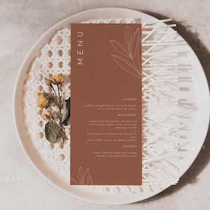 May include: A brown menu card with a white outline of leaves on the top and bottom. The text "MENU" is in white at the top of the card. The card is on a white plate with a woven pattern. The text on the card reads: "STARTERS", "Grilled asparagus, lemon, and red tomato pesto, and pan-fried tiger prawns", "MAIN COURSE", "Roasted duck leg glazed with herb and walnut crust, potato gratin and green asparagus bundles", "Grilled breast of chicken put with garlic, pearl barley risotto, and glazed chestnuts", "DESSERTS", "Fresh berries and yogurt with orange segments and honey bee sauce".