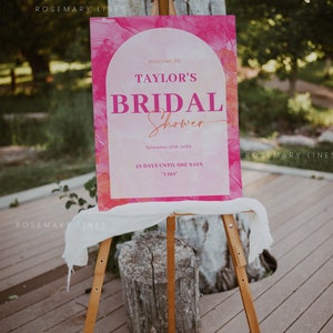May include: A pink and white bridal shower sign on a wooden easel. The sign reads "Welcome to Taylor's Bridal Shower" with the date and countdown to the wedding. The background has a watercolor effect.
