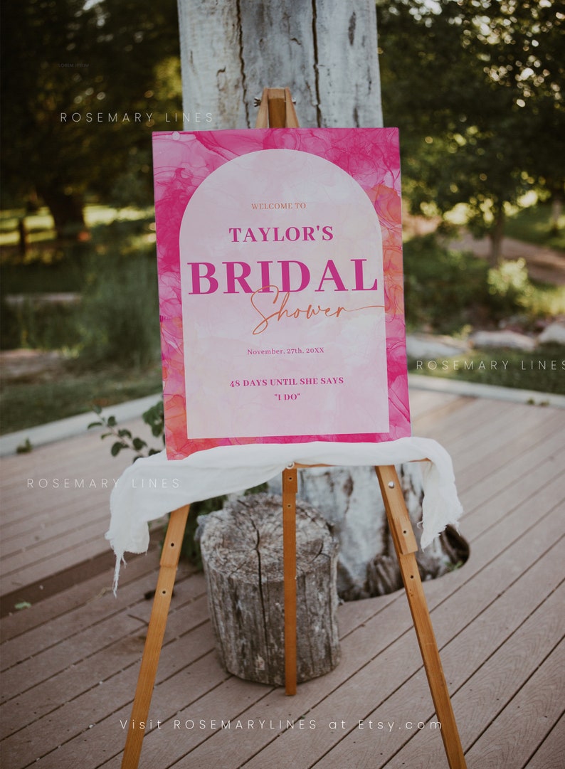 May include: A pink and white bridal shower sign on a wooden easel. The sign reads "Welcome to Taylor's Bridal Shower" with the date and countdown to the wedding. The background has a watercolor effect.