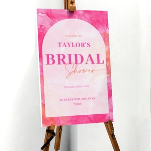 May include: A pink and white bridal shower sign on a wooden easel. The sign reads "Welcome to Taylor's Bridal Shower" with the date and "48 days until she says I do."