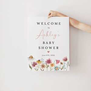 May include: A welcome sign for a baby shower with a floral border. The sign says "Welcome to Ashley's Baby Shower" with a pink heart and the date "June 27th, 2022".