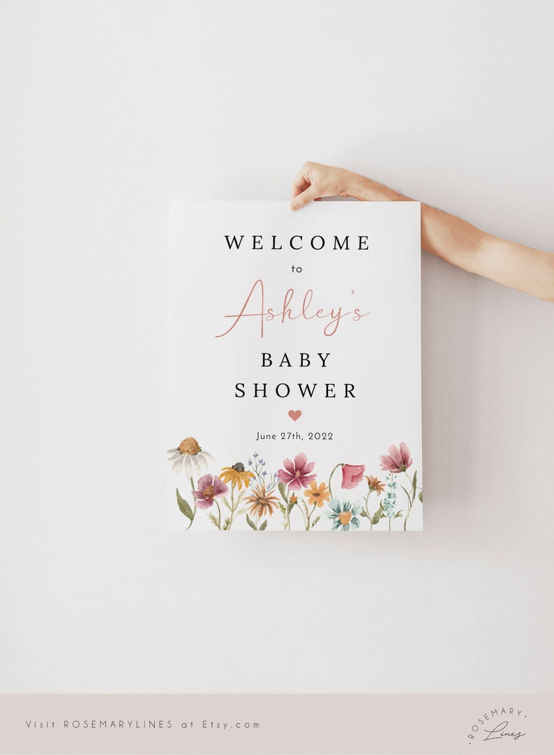 May include: A welcome sign for a baby shower with a floral border. The sign says "Welcome to Ashley's Baby Shower" with a pink heart and the date "June 27th, 2022".