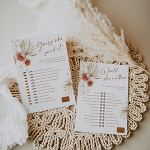 May include: Two printable bridal shower games, "Guess Who Said It" and "Would She Rather", with floral and botanical accents. The games are printed on white paper with brown text and decorative elements. The games are displayed on a woven placemat with dried flowers.
