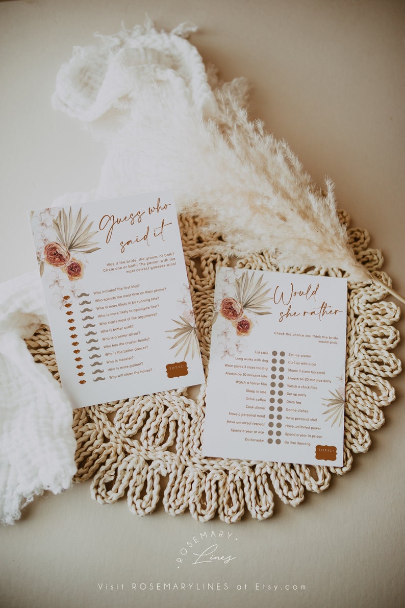 May include: Two printable bridal shower games, "Guess Who Said It" and "Would She Rather", with floral and botanical accents. The games are printed on white paper with brown text and decorative elements. The games are displayed on a woven placemat with dried flowers.
