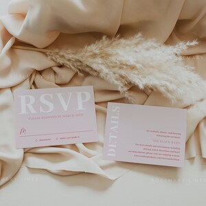 May include: Two pink wedding stationery cards with white text, set on a beige fabric. The RSVP card has a handwritten initial and response options. A sprig of dried pampas grass adds a natural touch to the scene.