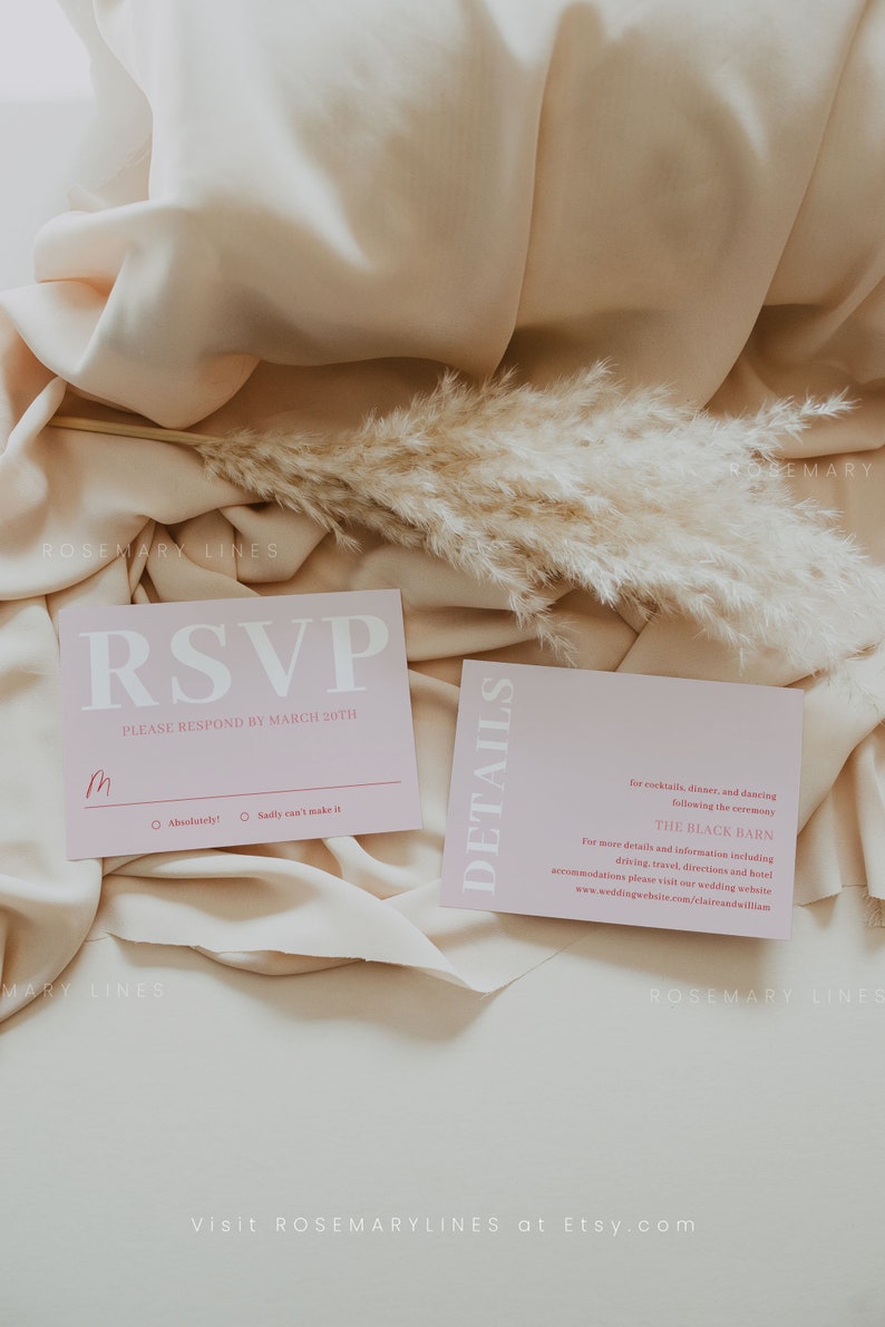 May include: Two pink wedding stationery cards with white text, set on a beige fabric. The RSVP card has a handwritten initial and response options. A sprig of dried pampas grass adds a natural touch to the scene.