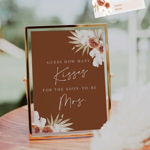 May include: A framed sign and a small card for a bridal shower game. The sign is 5 x 7 inches and features the text "Guess How Many Kisses" on a brown background, with floral accents. The card is 3.5 x 2 inches.